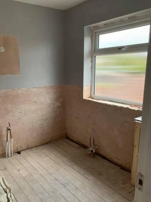 A bathroom stripped back to bare plaster, with a window and pipework showing