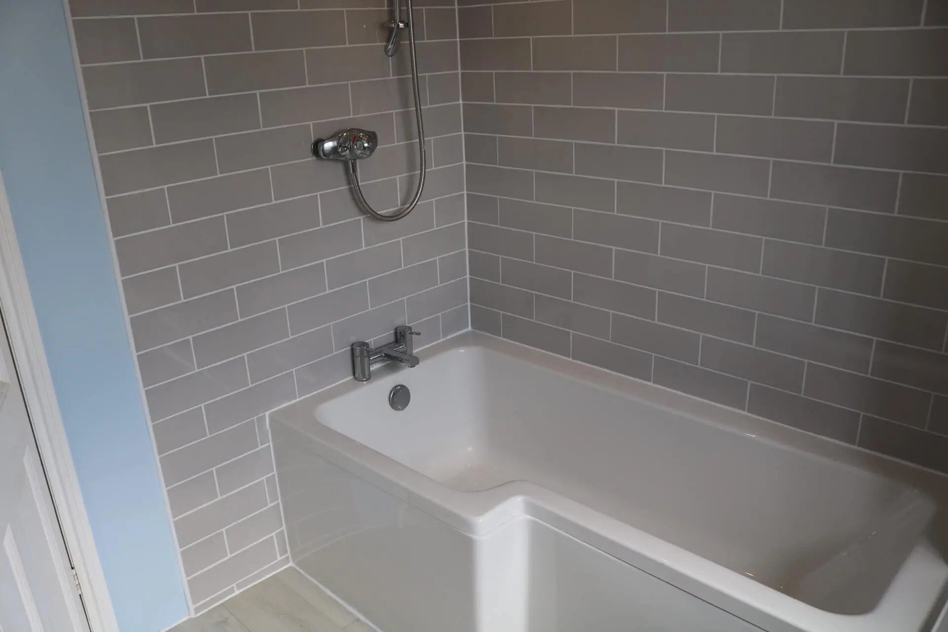 A white bath with a shower over it and grey metro tiles on the walls