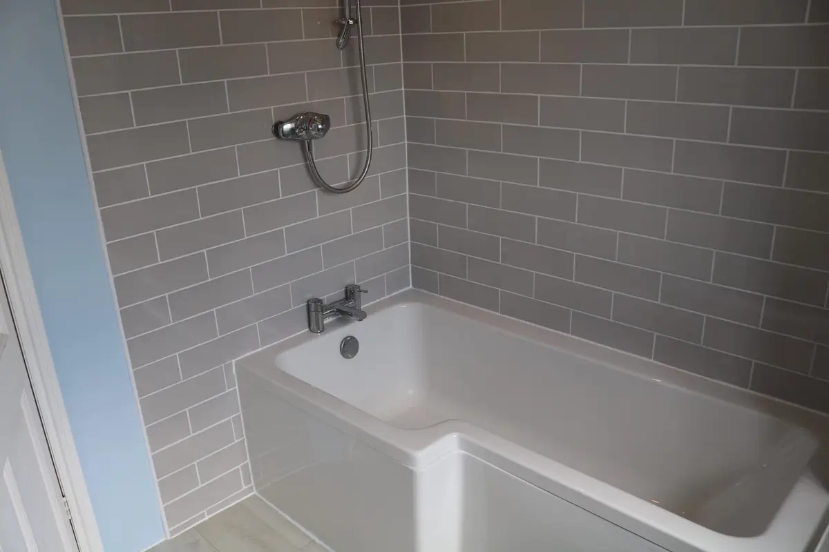A white bath with a shower over it and grey metro tiles on the walls